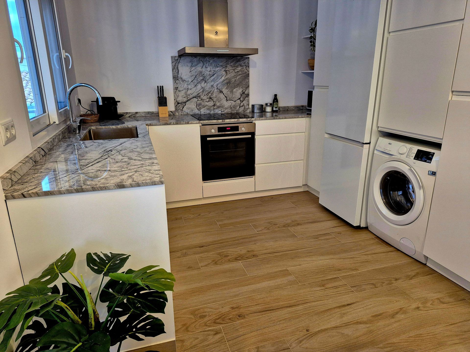 Cocina moderna con gabinetes blancos, encimeras de granito y pisos imitación madera. Incluye electrodomésticos y una maceta.