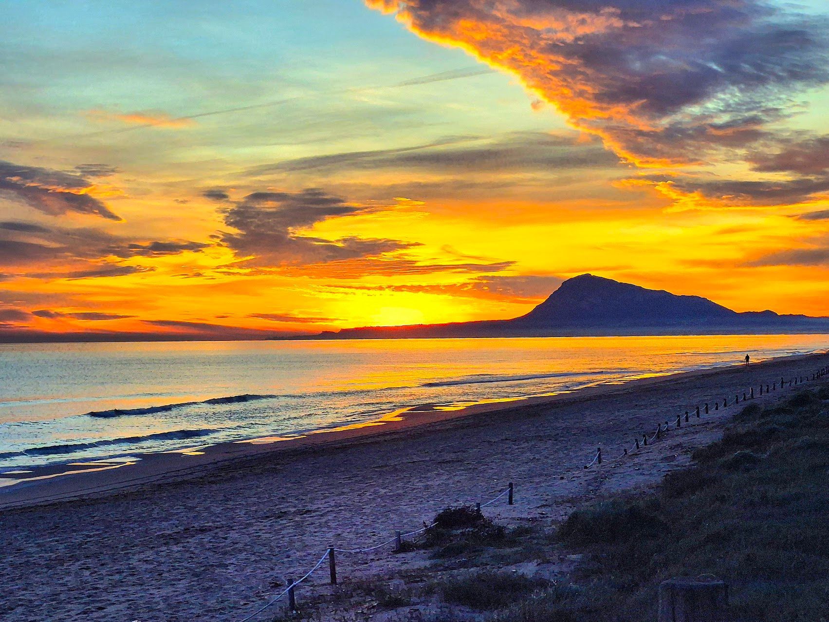 Atardecer en una playa de arena oscura con Gibraltar a lo lejos. El cielo naranja y amarillo se refleja en el agua.