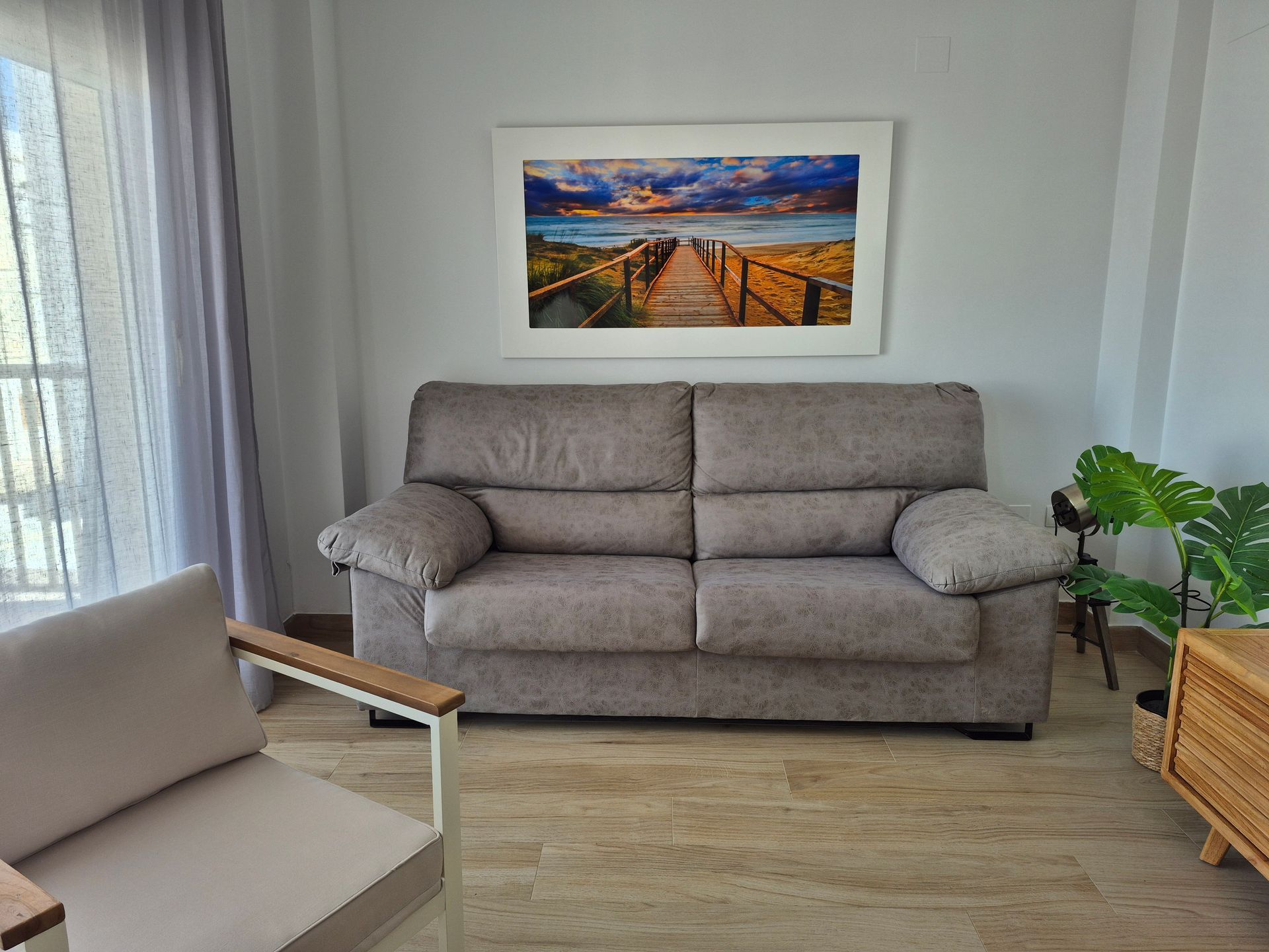 Luminoso salón con sofá gris, cuadro paisajístico enmarcado y macetero de mimbre a la derecha.
