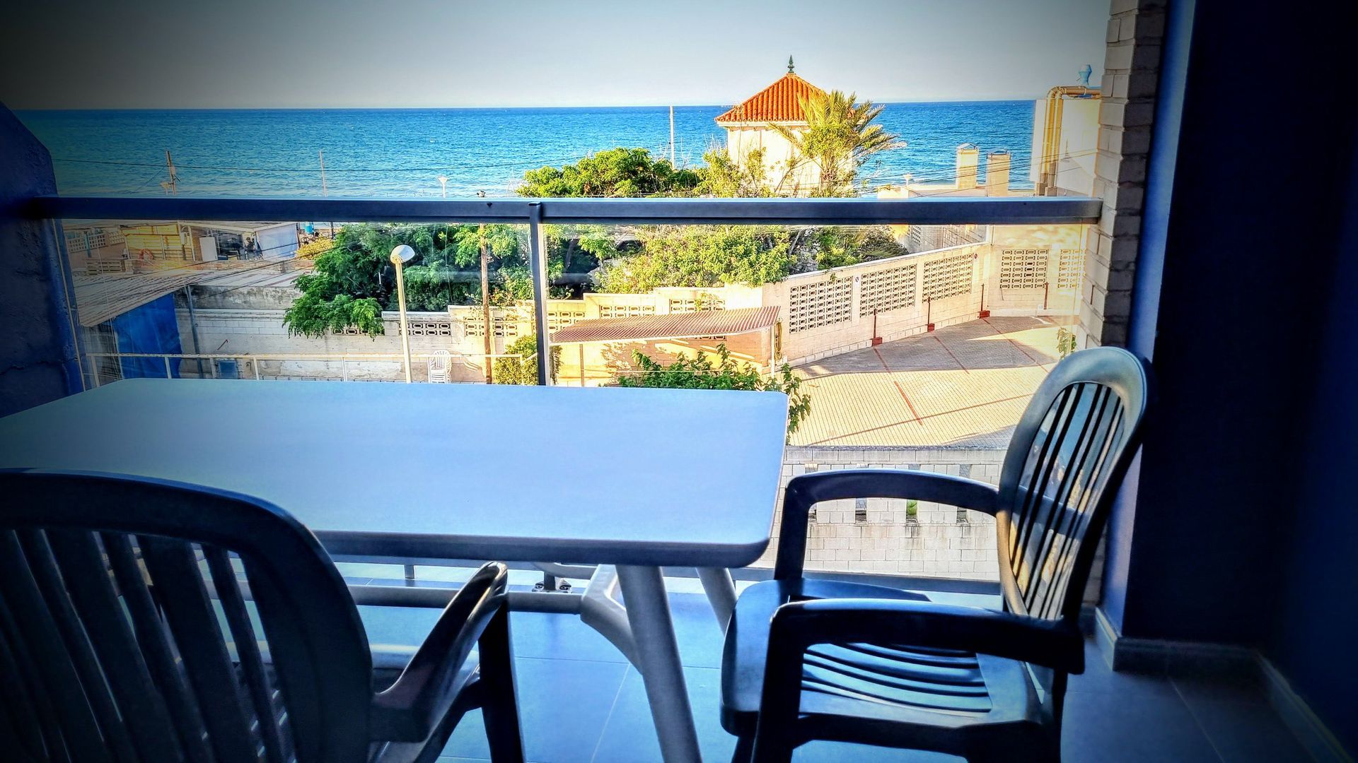 Un balcón junto al mar con mesa y sillas, con vista al océano y a un edificio.