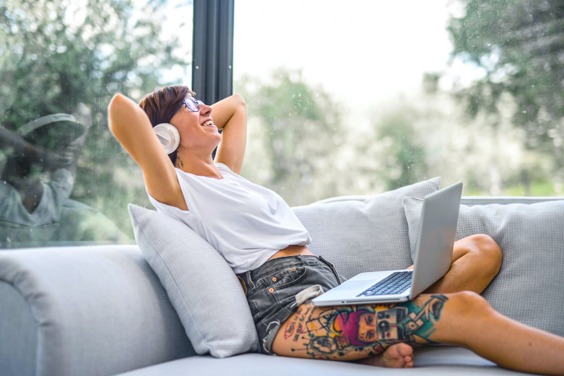 Woman wearing headphones relaxes on a couch with a laptop, laughing, large leg tattoo visible.
