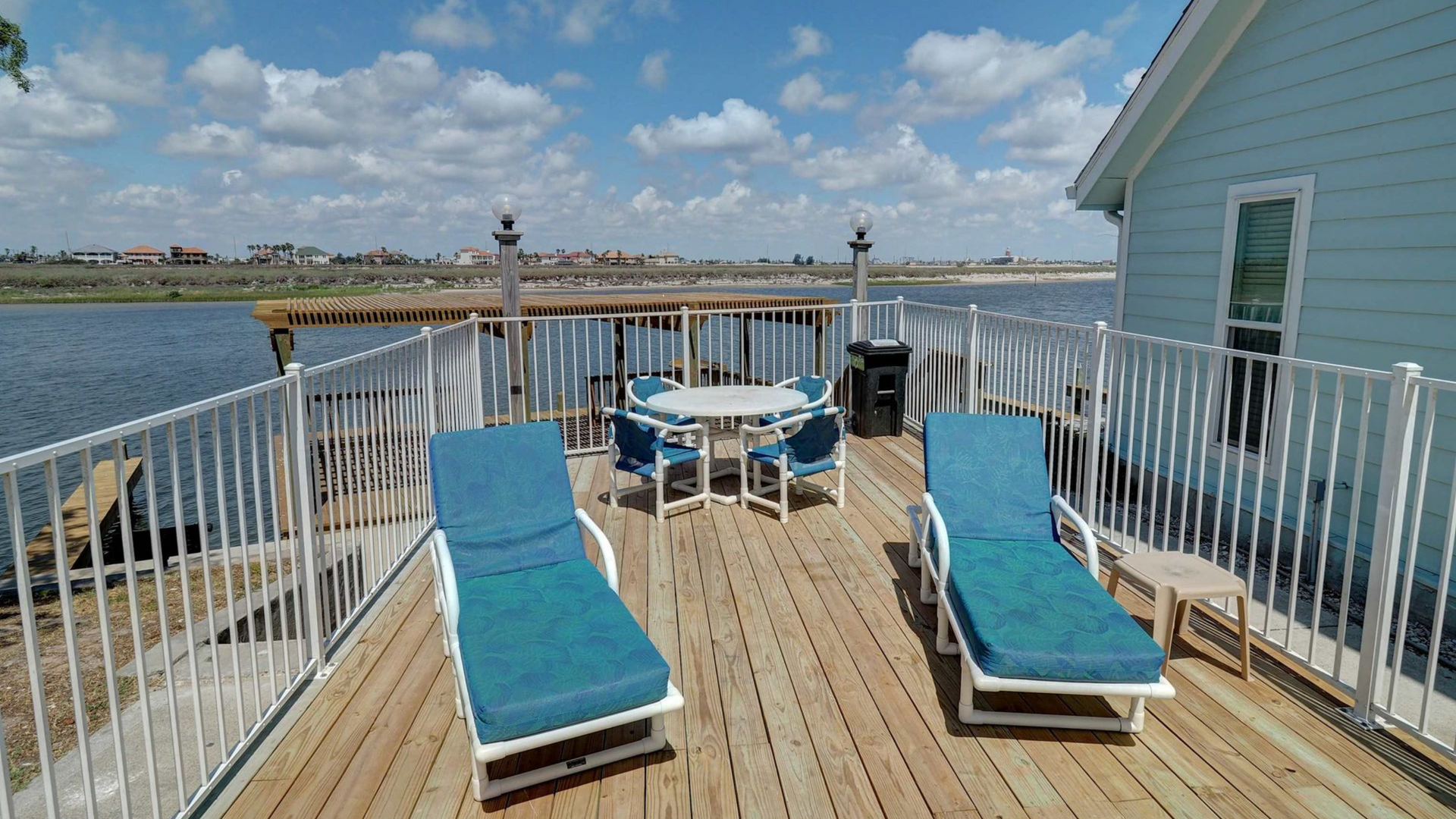 Waterfront deck with two turquoise lounge chairs, a table, and railing beside a house under a blue sky