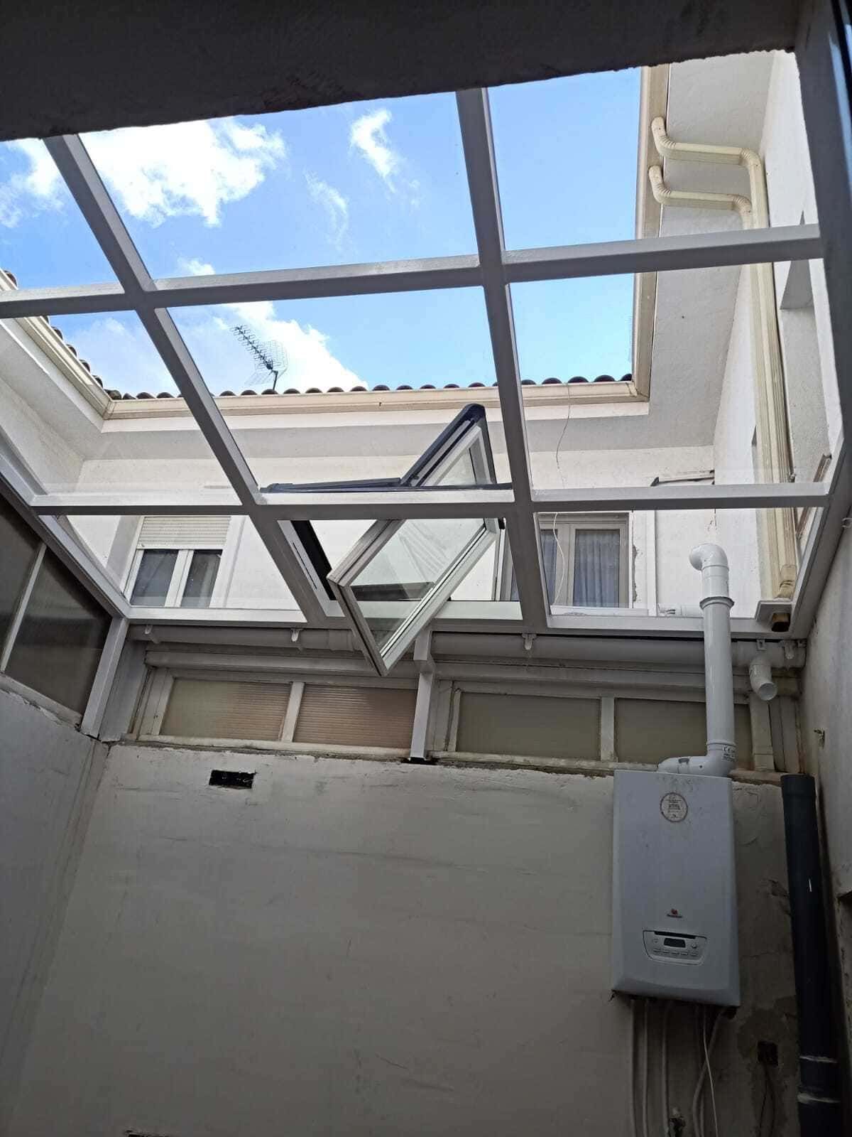 Patio interior con techo de cristal abierto, paredes blancas y una caja eléctrica a la derecha.