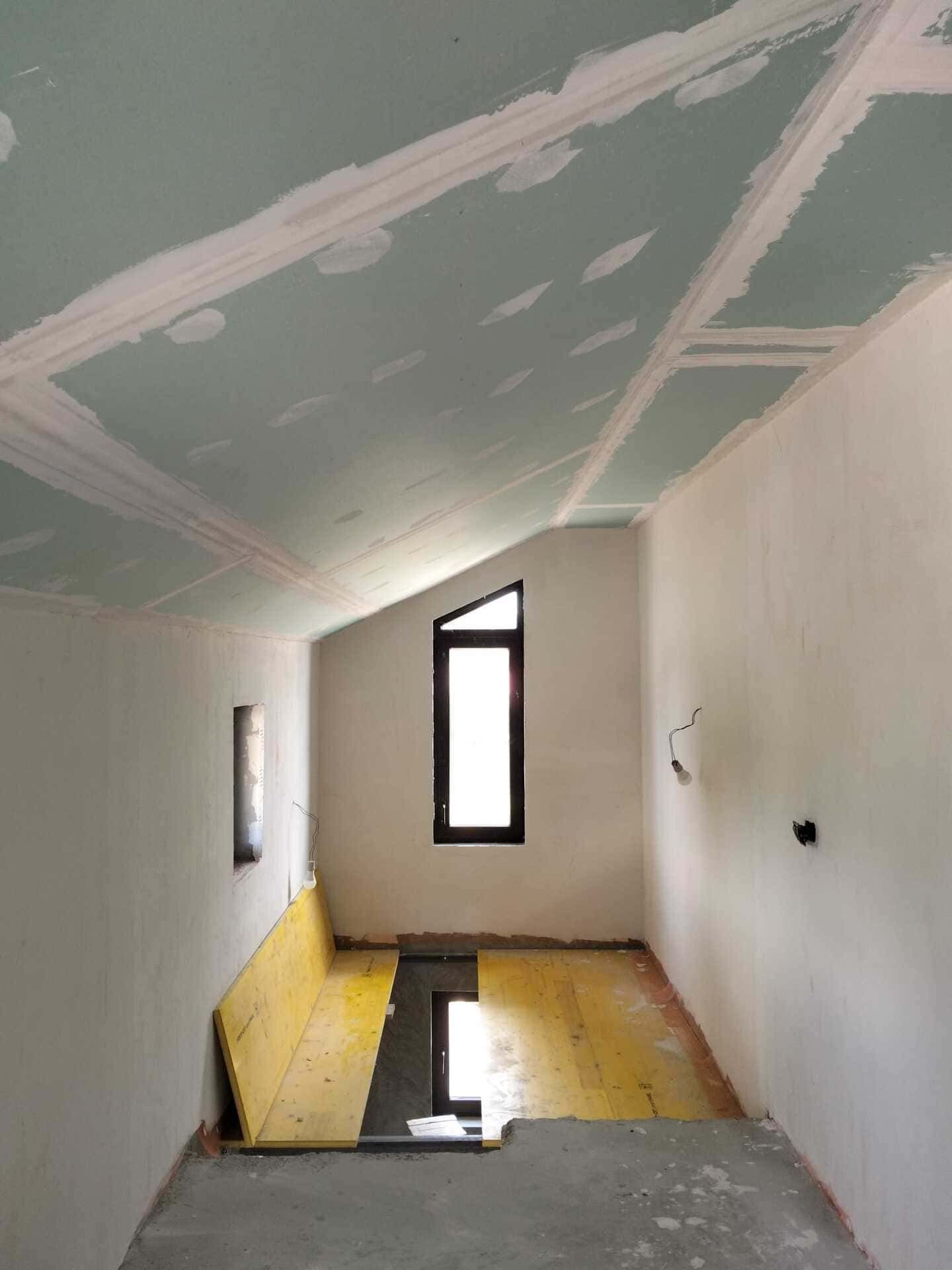 Habitación estrecha y sin terminar con paredes blancas, techo inclinado y una pequeña ventana, reflejada en un agujero en el suelo inundado.