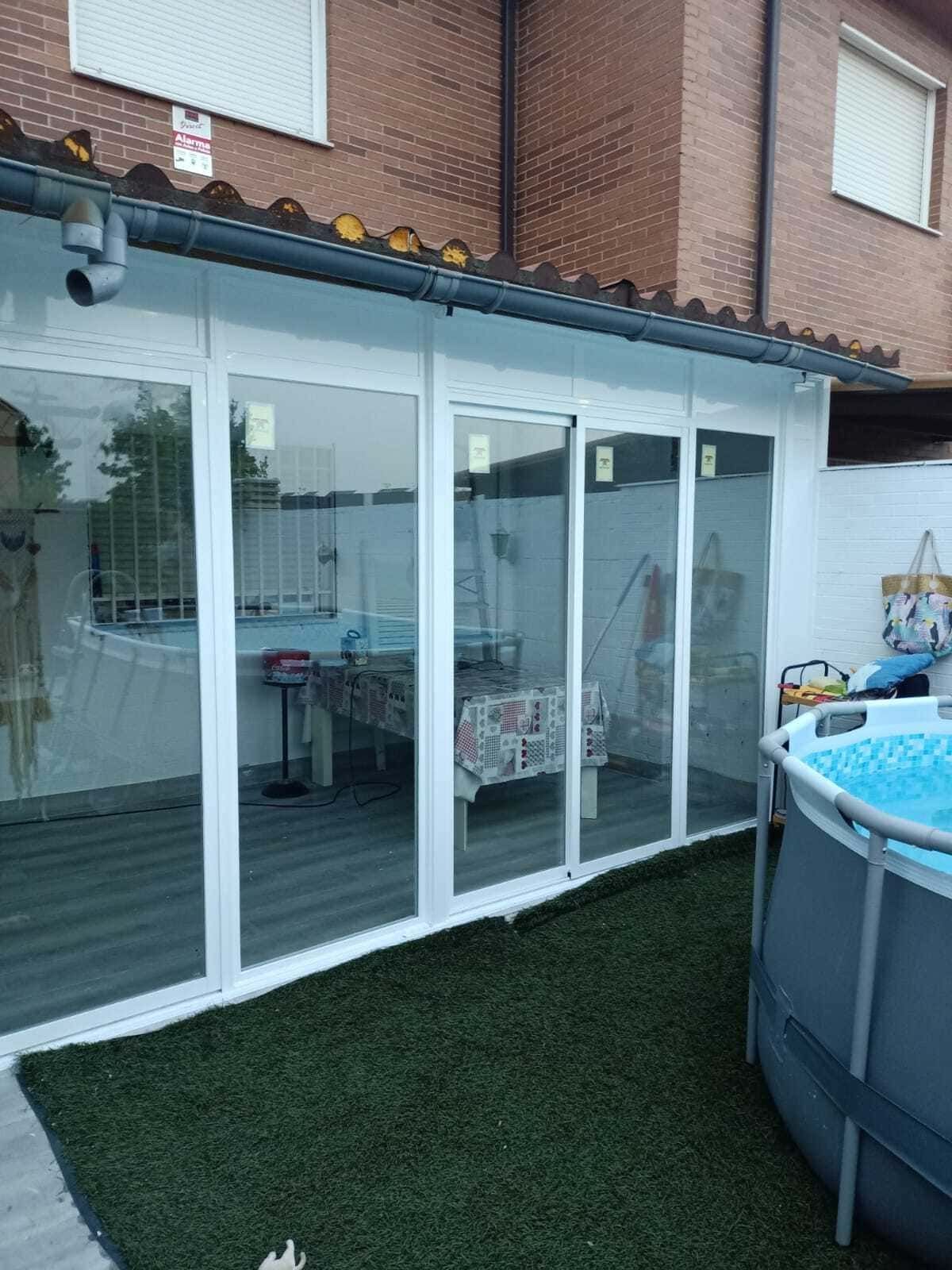 Terraza acristalada en el patio trasero con puertas corredizas de vidrio y una piscina elevada azul junto a césped artificial.