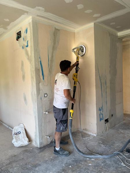 Un trabajador lija una pared interior con una lijadora de pértiga y una manguera de aspiradora durante una reforma.