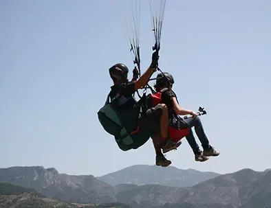 Un parapente biplaza con dos personas volando contra un cielo despejado sobre montañas onduladas.