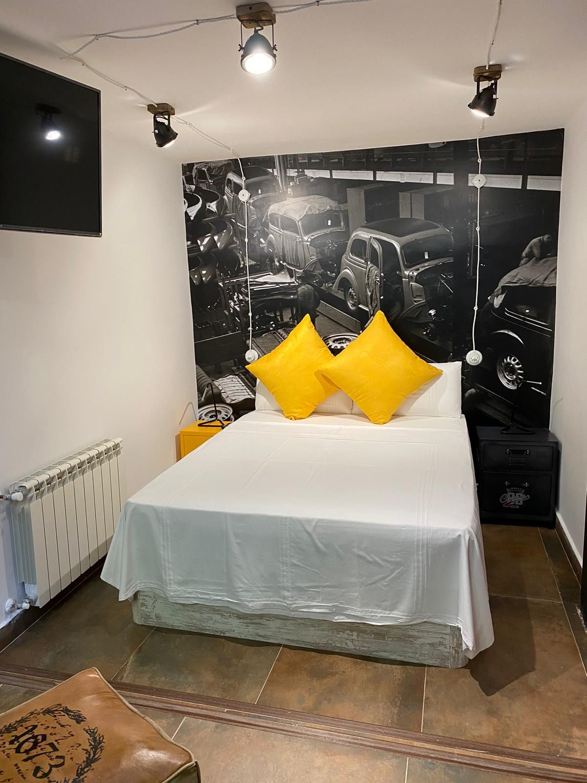 Dormitorio con una cama, almohadas amarillas y un mural en blanco y negro con temática de automóviles.