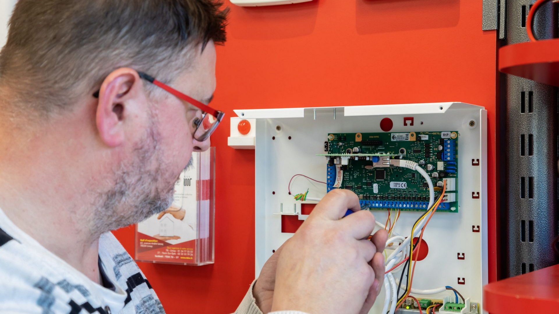 Un homme portant des lunettes effectue des branchements électriques à l'intérieur d'un boîtier de commande blanc, sur un mur rouge.