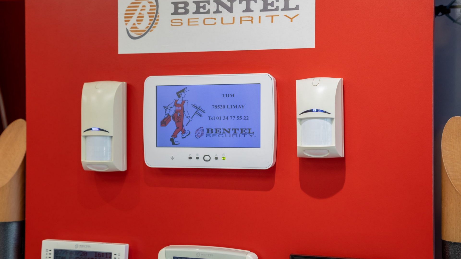 Présentation d'équipements de sécurité Bentel sur un mur rouge, comprenant un panneau de commande et des détecteurs de mouvement.
