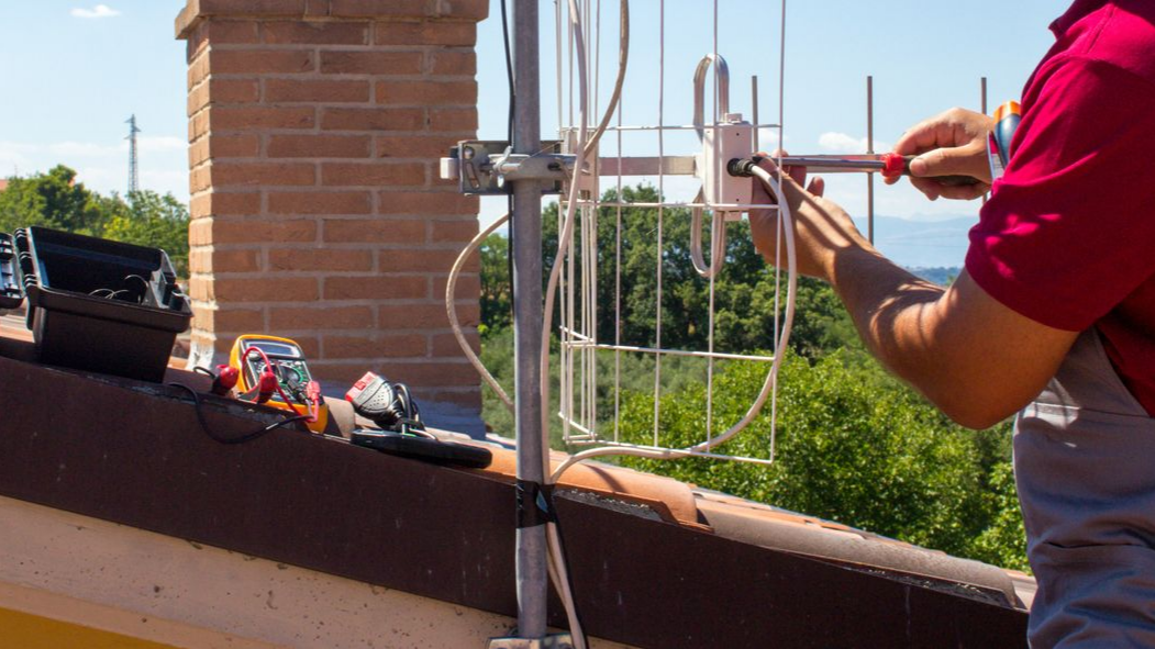 Un technicien sur un toit ajuste une antenne avec un tournevis.