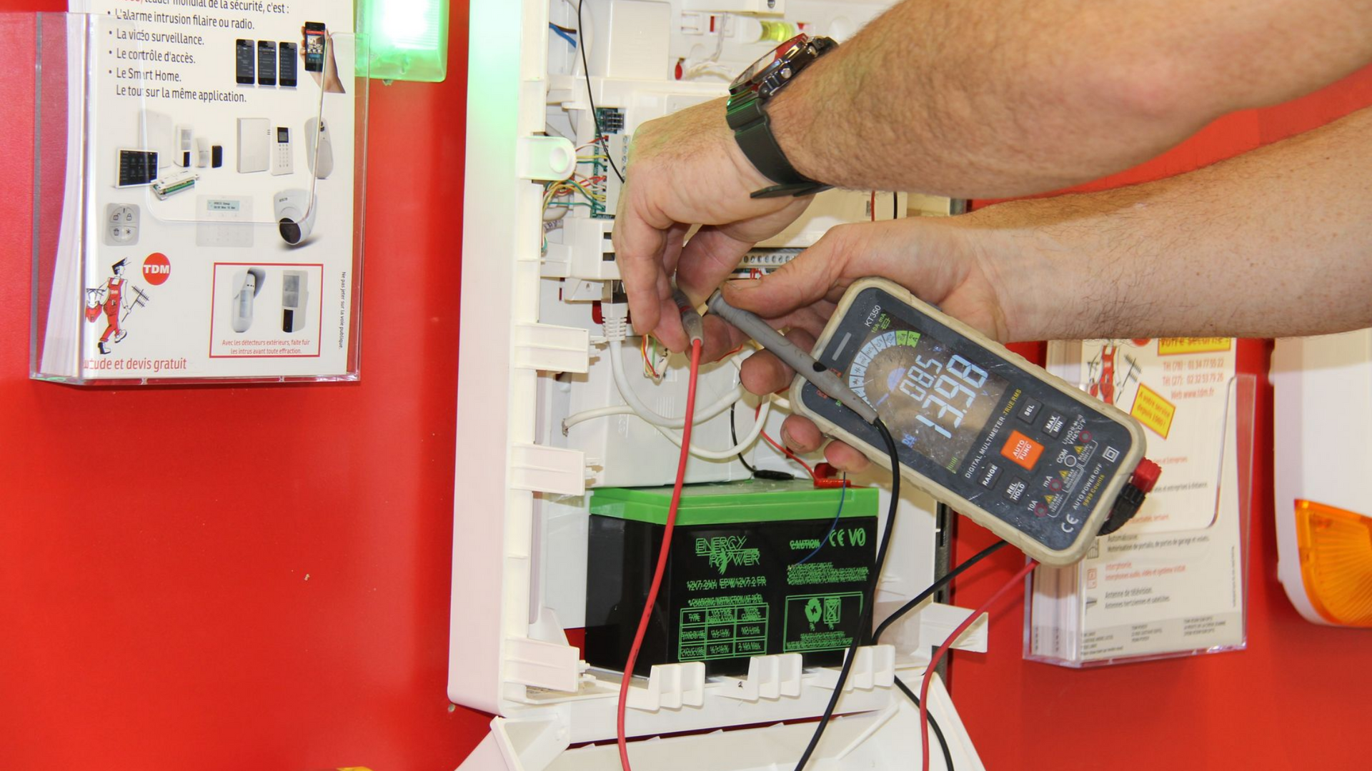 Un technicien vérifie des branchements électriques à l'intérieur d'un boîtier de commande blanc, sur un mur rouge.