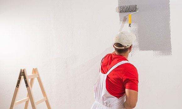 artisan avec une casquette et un t-shirt rouge qui applique de la peinture grise sur un mur blanc