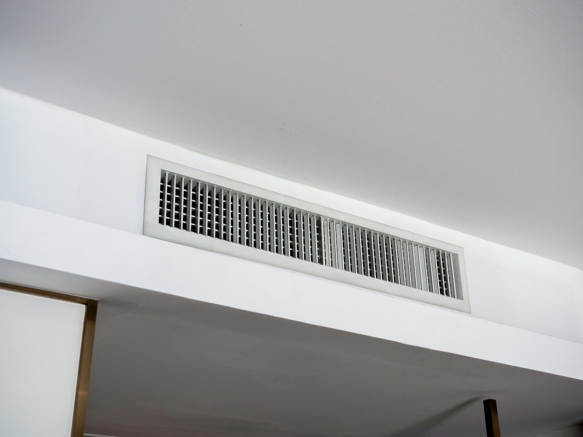 Grille d'aération blanche au plafond, fixée sur un mur au-dessus d'une porte, laissant apparaître une partie de l'encadrement en dessous.