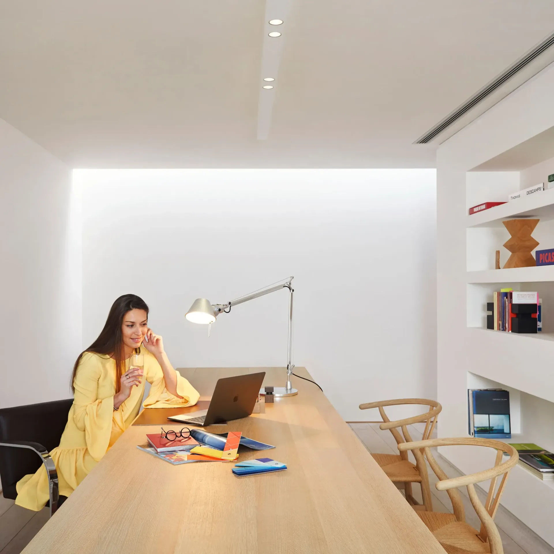 Une femme travaille à un bureau en bois dans un bureau moderne et lumineux, avec un ordinateur portable et une lampe de bureau à proximité.