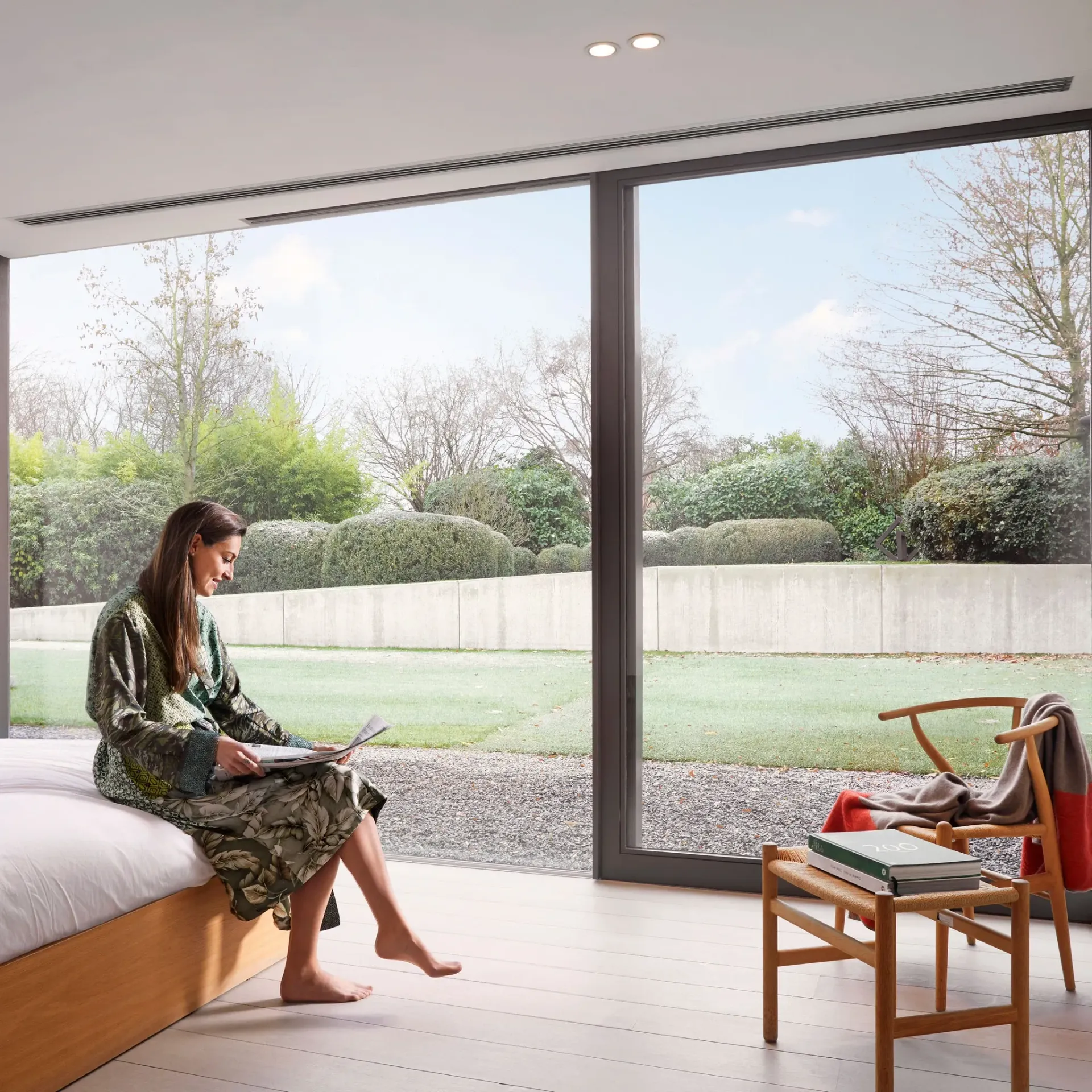 Une personne assise sur un lit dans une chambre moderne, près de grandes fenêtres, regarde un ordinateur portable avec vue sur le jardin.
