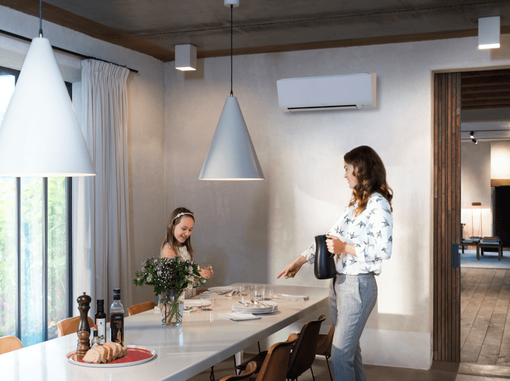 Deux femmes dressent la table dans une cuisine moderne et lumineuse, éclairée par des suspensions et agrémentée d'un vase de fleurs.