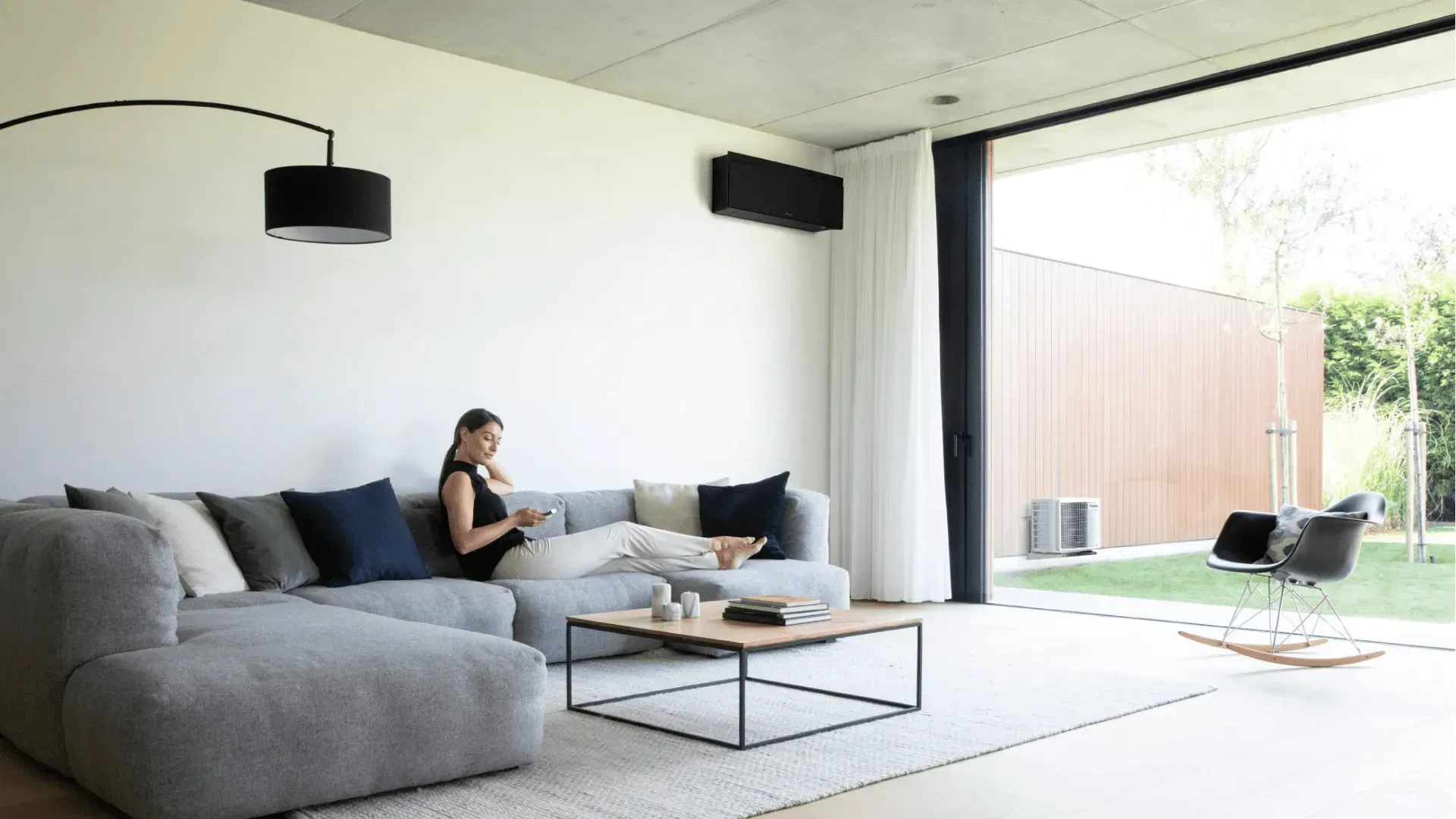 Salon moderne avec canapé d'angle gris, suspensions lumineuses noires et vue dégagée sur une terrasse ensoleillée.