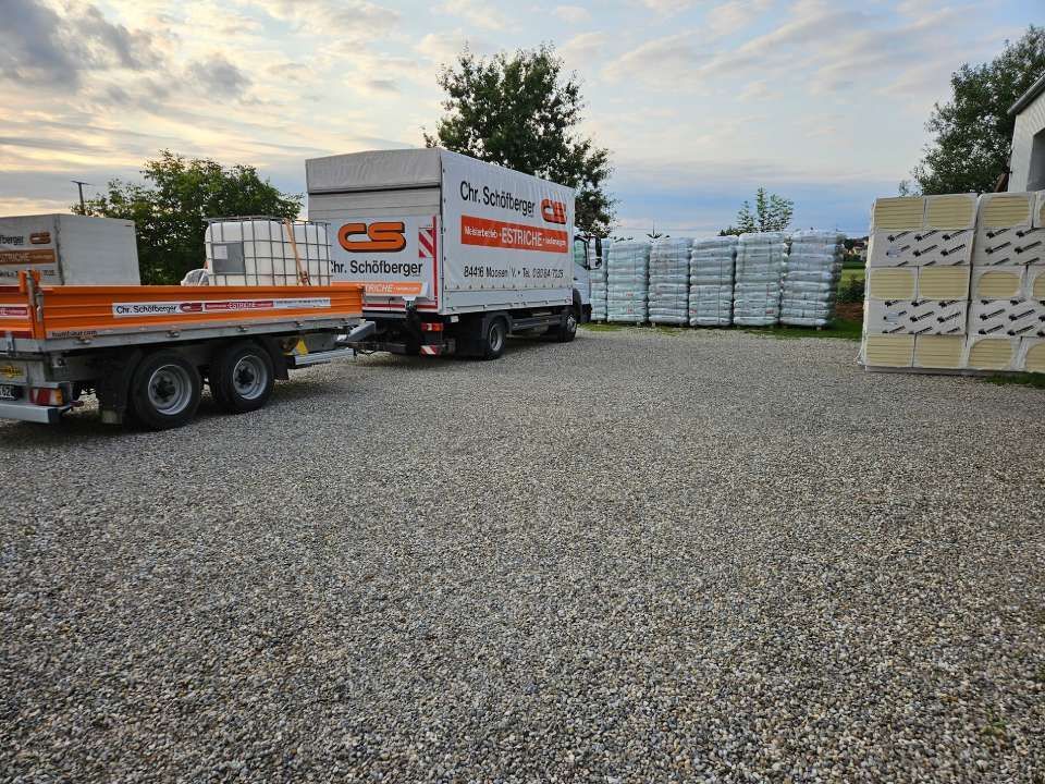 LKW und Anhänger auf Schotter mit Stapeln von Baumaterialien auf einem Parkplatz.