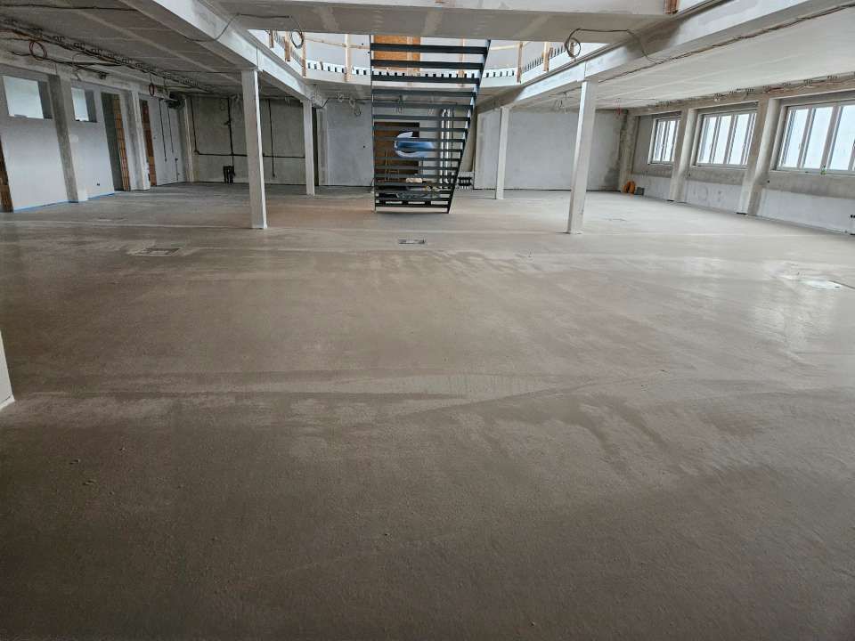 Ein leerer, unfertiger Industrieraum mit Betonboden, Säulen, Fenstern und einer Treppe, die nach oben führt.