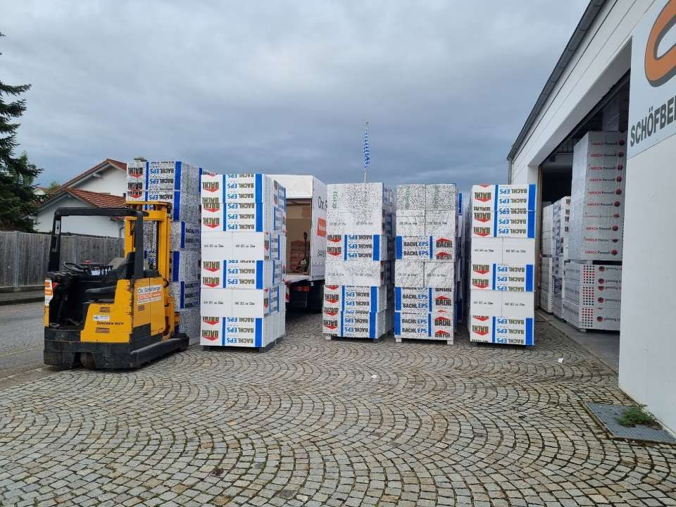 Gelber Gabelstapler und Paletten mit Kisten vor einem Gebäude, bewölkter Himmel.