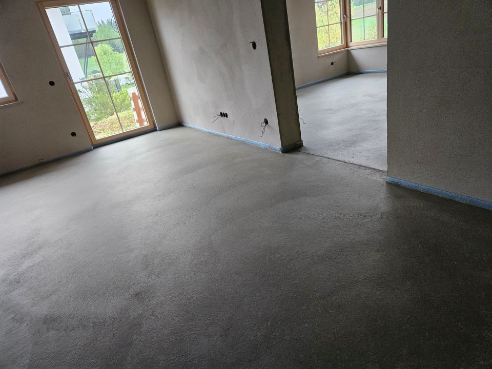 Innenansicht eines Zimmers mit grauem Betonboden, Fenstern und hellen Wänden.