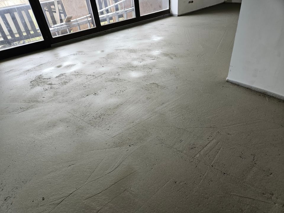 Grauer Betonboden mit sichtbaren Markierungen und Lichtreflexionen, neben einem Fenster und einer weißen Wand.