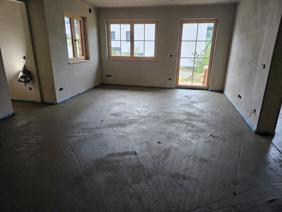 Innenansicht eines im Bau befindlichen Raumes mit unfertigen Wänden und Betonboden, Fenstern und einer Türöffnung.