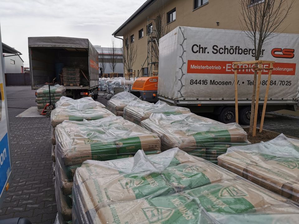 Im Freien stehen neben zwei Lastwagen Stapel verpackter Baumaterialien. Ein Lastwagen ist mit einer Plane abgedeckt, der andere trägt ein aufgedrucktes Logo.