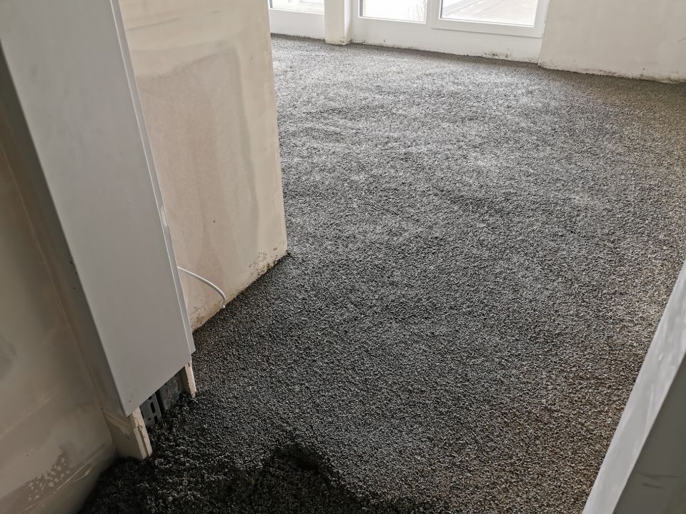 Zimmer mit dunkelgrauem, körnigem Bodenbelag. Teilweise fertiggestellter Innenbereich mit weißen Zierleisten und Türrahmen.