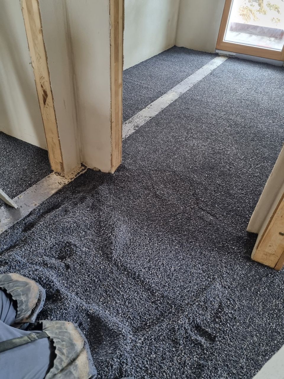 Der Innenraum ist mit dunkelgrauen, losen Granulaten bedeckt und befindet sich neben einer Türöffnung und einem Holzrahmen.