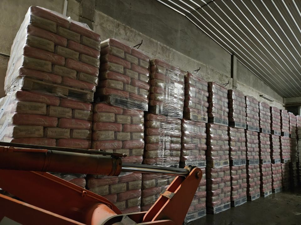 In einem Lagerhaus stapeln sich Säcke mit Material, vermutlich Zement oder Ähnliches. Im Vordergrund ist ein orangefarbener Gabelstaplerarm zu sehen.