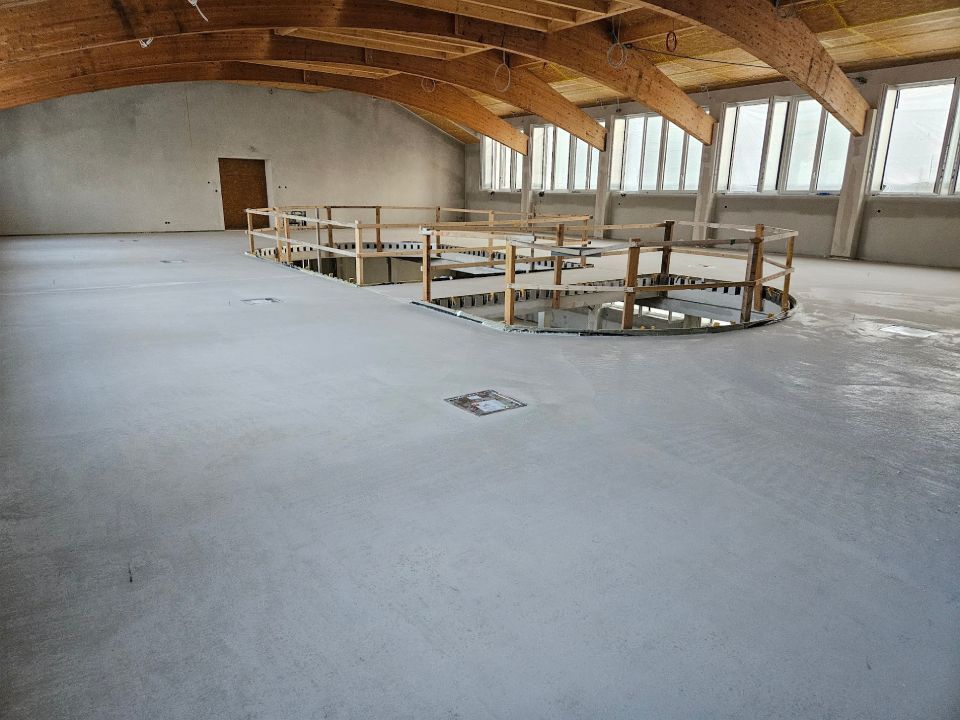 Innenansicht eines großen, unfertigen Gebäudes mit Betonboden und hölzernem Bogendach.