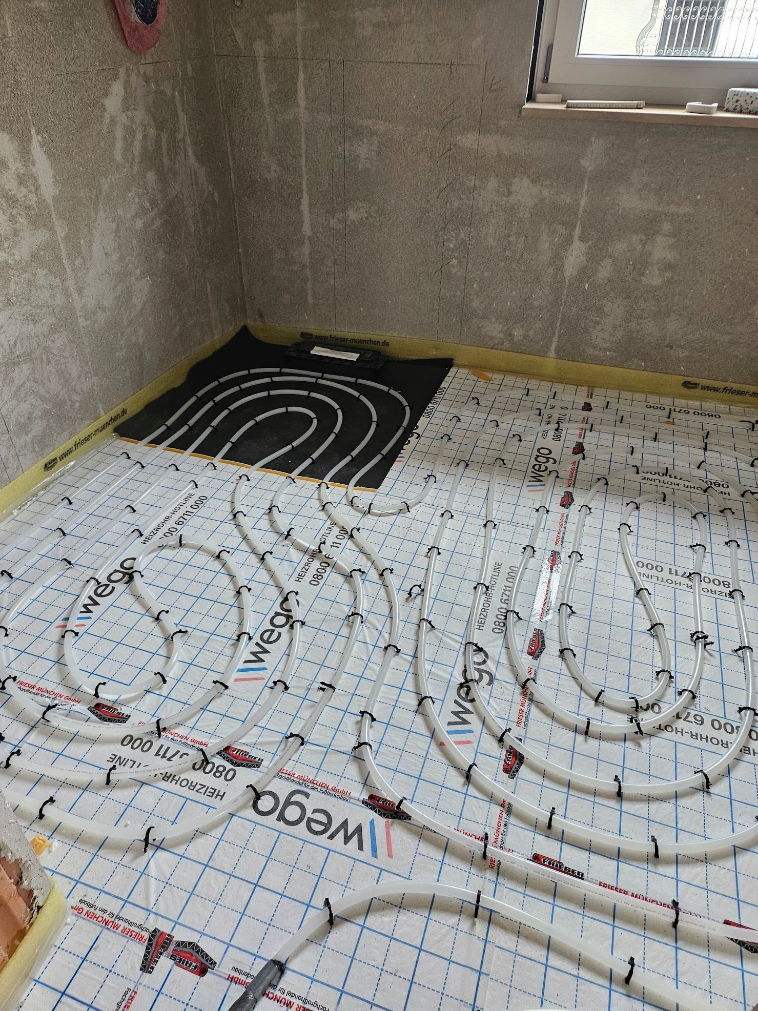 Weiße Rohre für eine Fußbodenheizung, verlegt auf einem Raster, in einem Raum mit unfertigen Betonwänden.