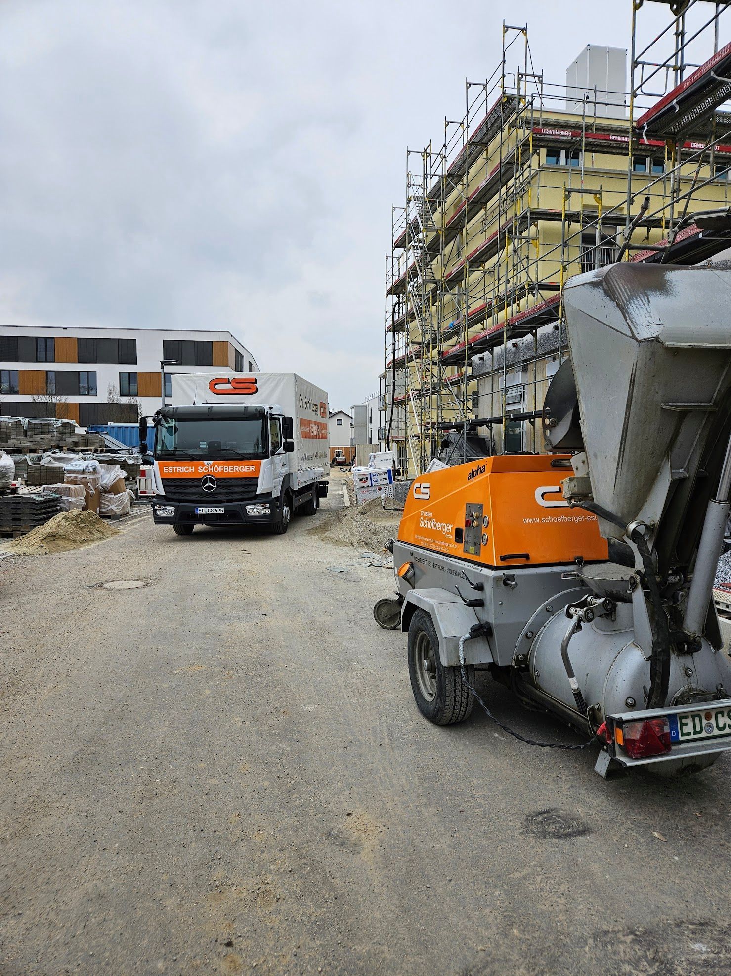 Baustelle mit einem LKW und Baumaschinen in der Nähe eines eingerüsteten Gebäudes, bewölkter Himmel.