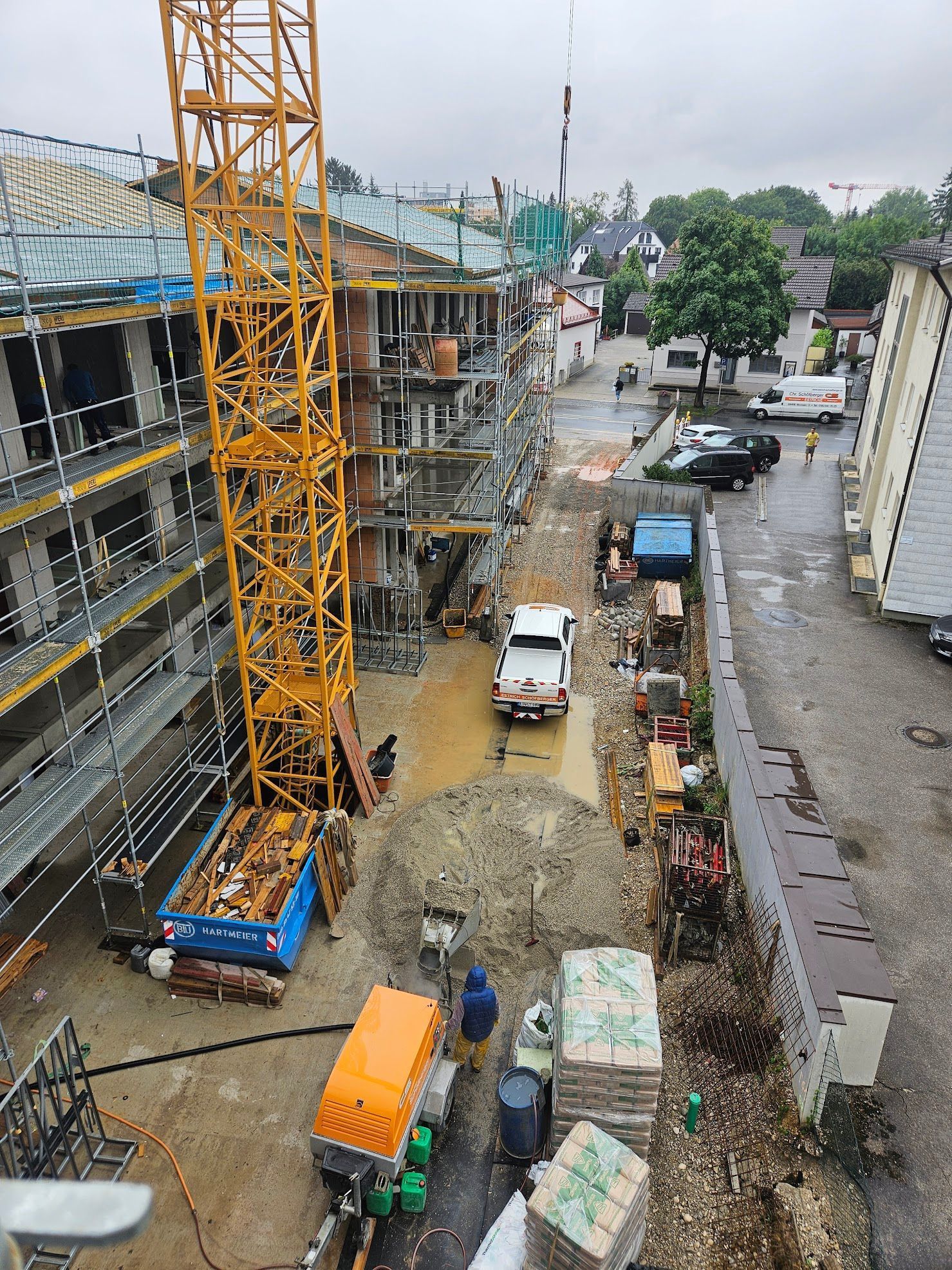 Baustelle mit gelbem Kran, Gebäude, LKW und Baumaschinen. Nasser, bewölkter Tag.