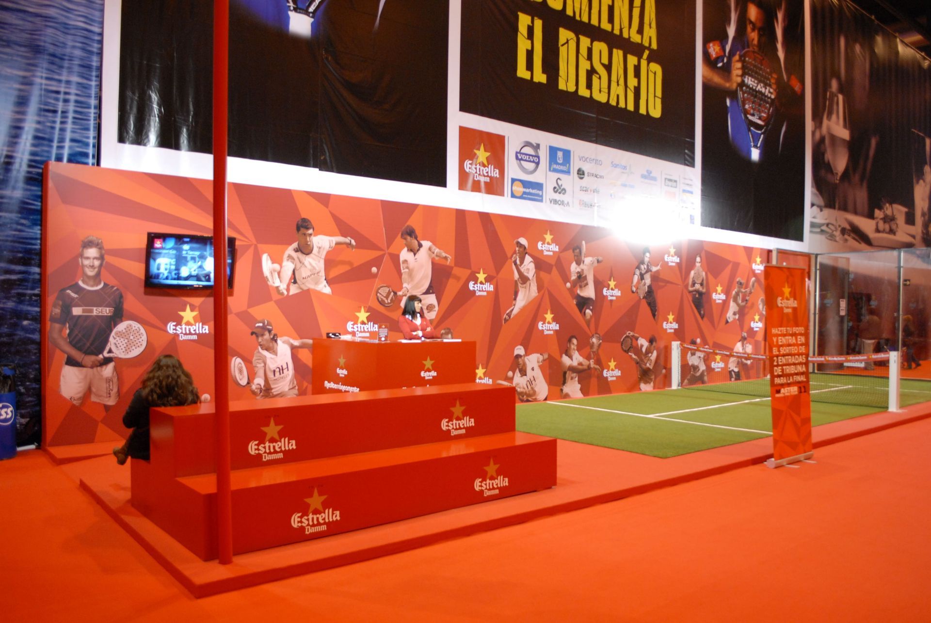 Exhibición de evento deportivo en color rojo y naranja con trofeo, imágenes de jugadores de fútbol, ​​césped y el logotipo de Estrella.