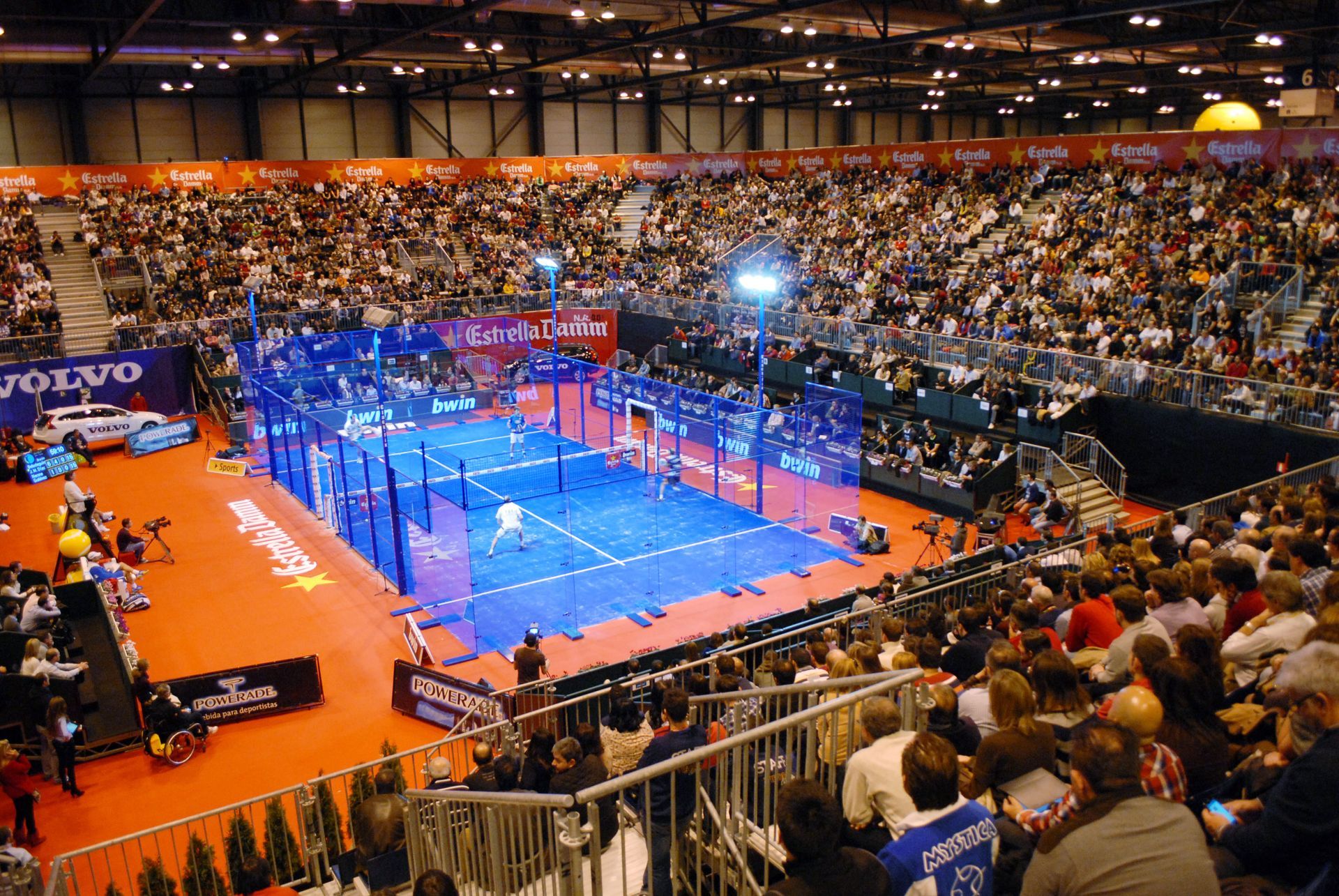 Partido de pádel en un estadio. Jugadores en la pista, rodeados de paredes de cristal. Un gran público observa.