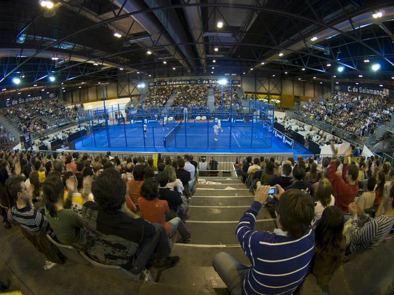 Pista de pádel cubierta llena de espectadores viendo un partido. Pista azul, luces brillantes, gente sentada.