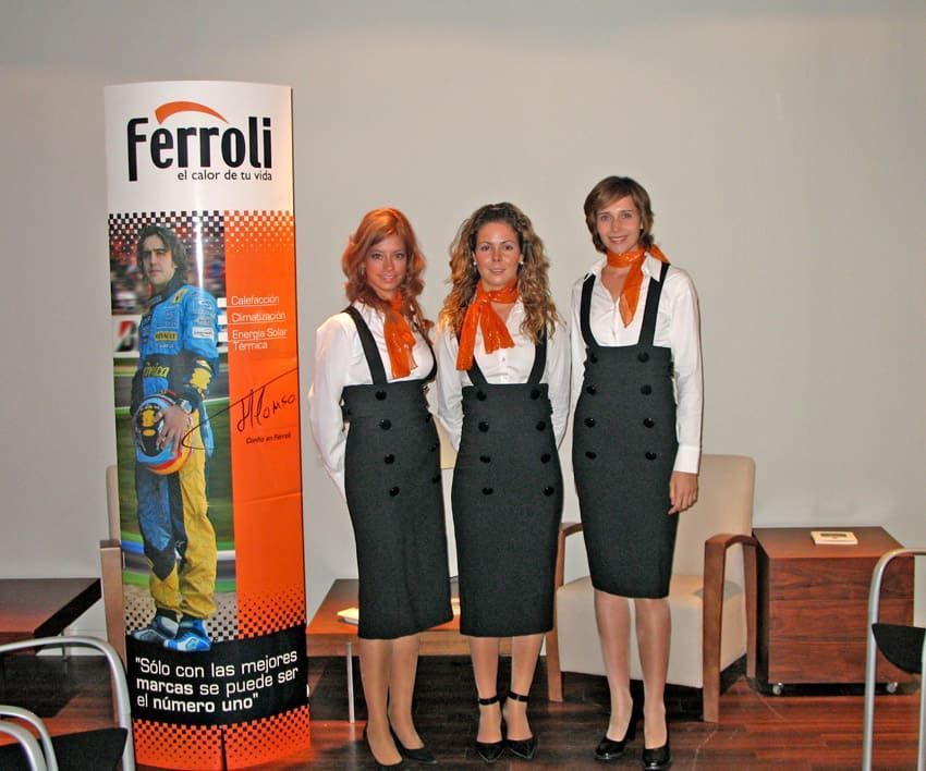 Tres mujeres con atuendos iguales posan junto a un anuncio de Ferroli. Llevan faldas negras, camisas blancas y pañuelos naranjas.