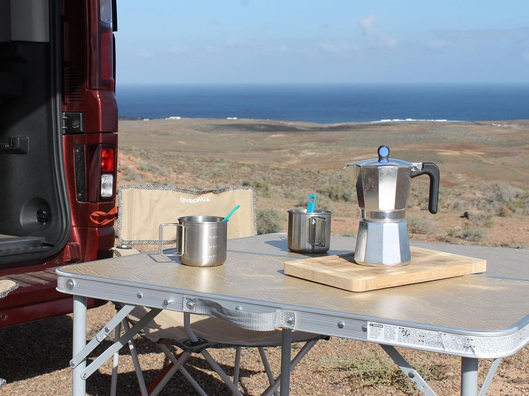 Una mesa con una cafetera y tazas encima delante de una camioneta roja.