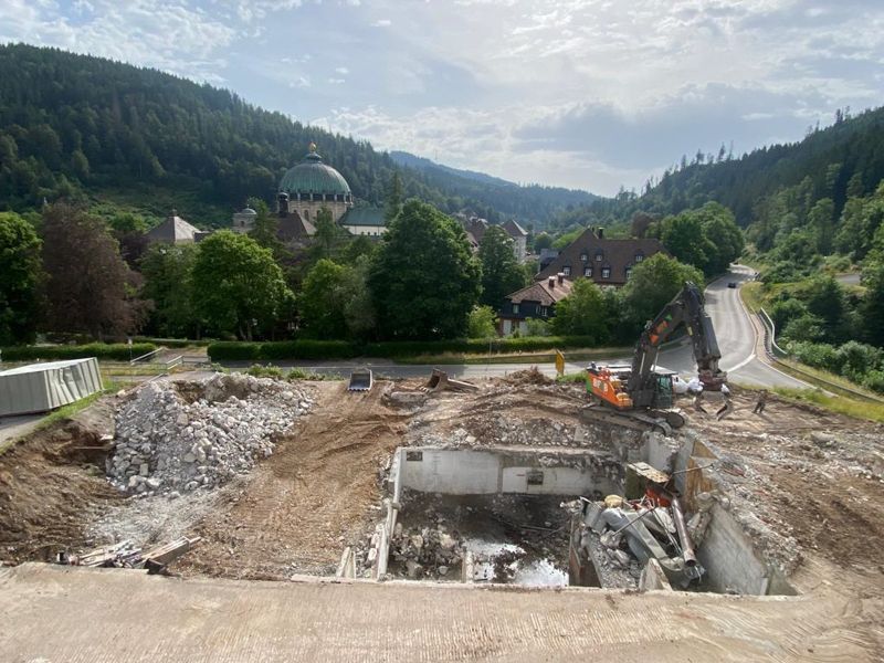 Baustelle mit einem Bagger, einem teilweise abgerissenen Gebäude und in einem Tal eingebetteten Gebäuden.