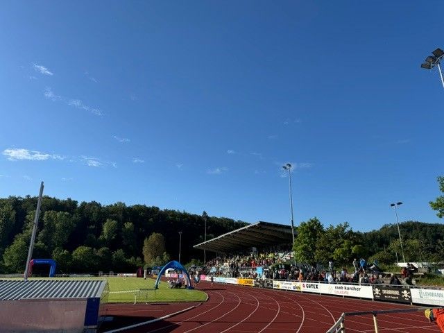 Outdoor running track with athletes, a covered grandstand, and a clear blue sky at a sports field