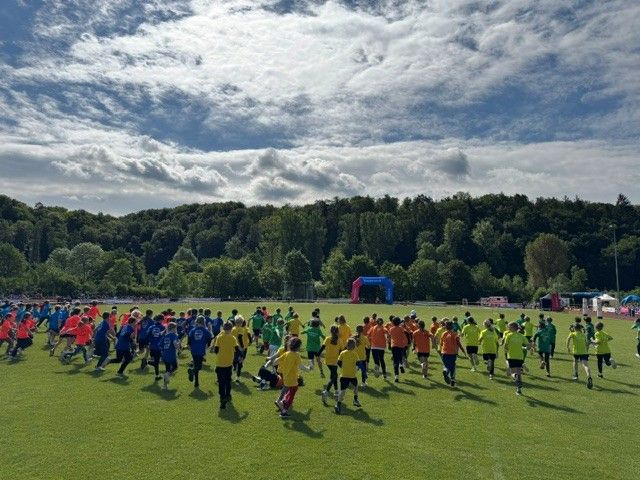 Youth soccer players in red, blue, yellow, and orange running on a grassy field under a cloudy sky