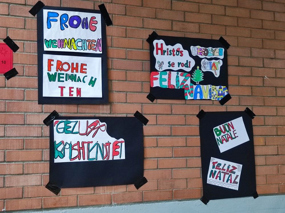 Festtagsgrüße in verschiedenen Sprachen auf einer Backsteinmauer: Deutsch, Spanisch und Italienisch.