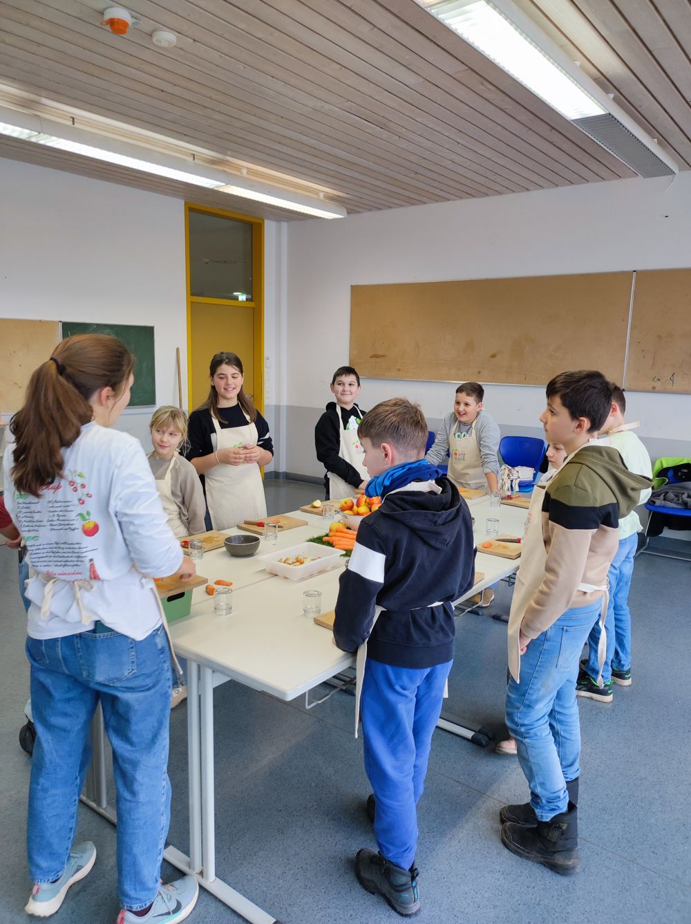 Kinder und Erwachsene backen in einem Klassenzimmer. Sie tragen Schürzen und versammeln sich um einen Tisch mit Essen.