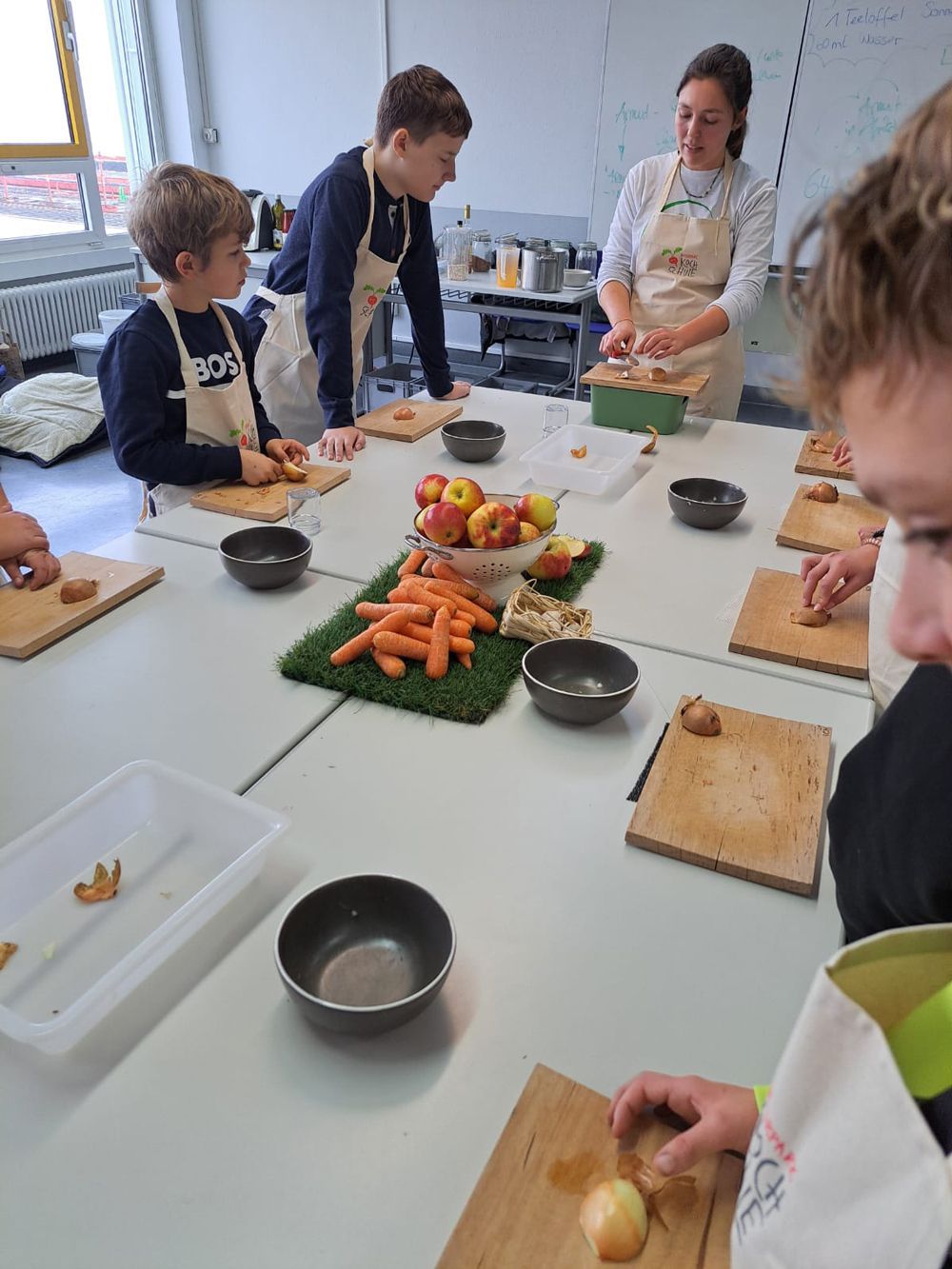 Kinder und ein Erwachsener in einer Küche, die an einem großen Tisch Essen zubereiten.