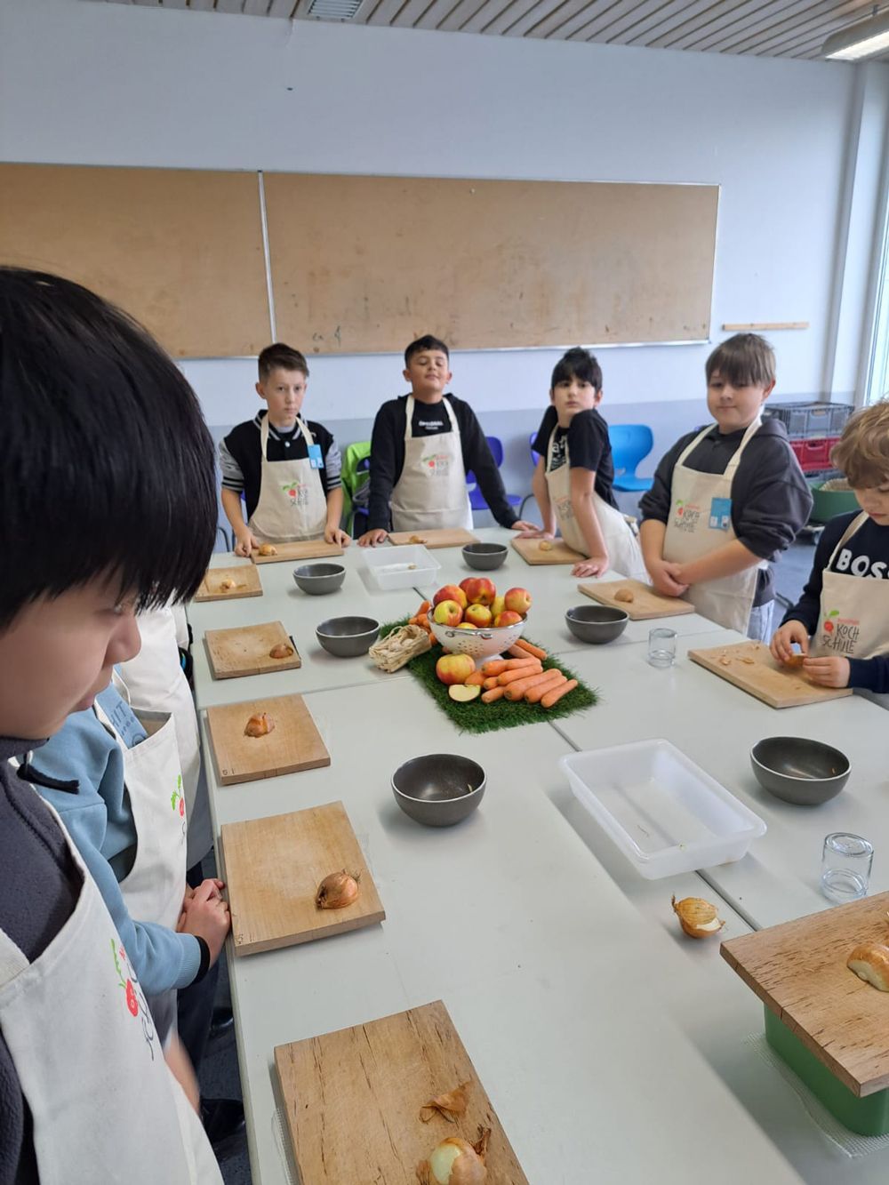 Kinder in Schürzen bereiten in einem Kochkurs an einem langen Tisch Speisen zu.