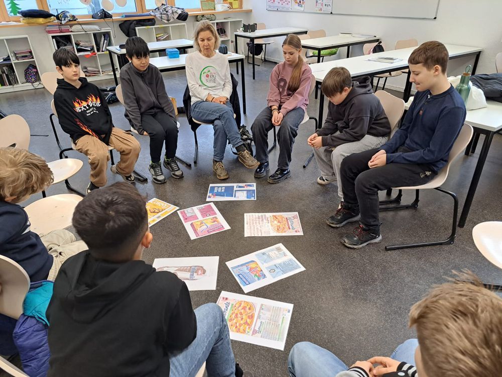 Eine Gruppe von Menschen sitzt im Kreis und betrachtet Bilder auf dem Boden. Klassenzimmersituation.