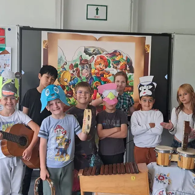 Kinder mit Musikinstrumenten und Hüten posieren in einem Klassenzimmer vor einem farbenfrohen Hintergrund.